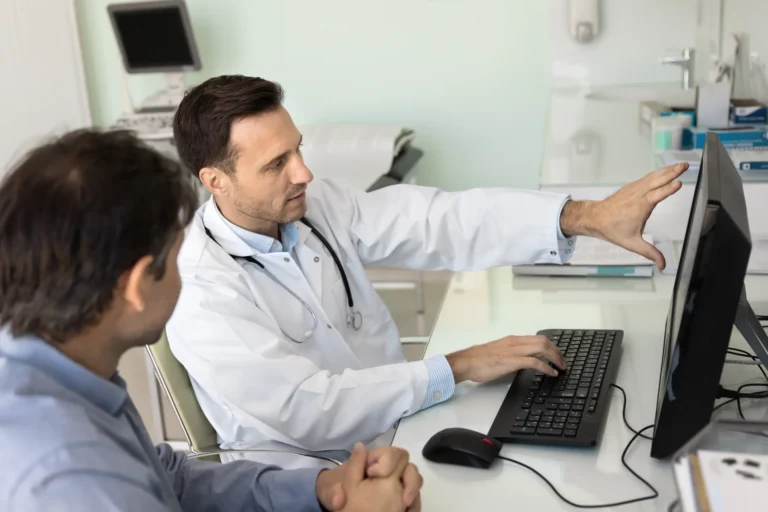 A doctor in a white coat points at a computer monitor while discussing information with a patient seated beside him in an office.