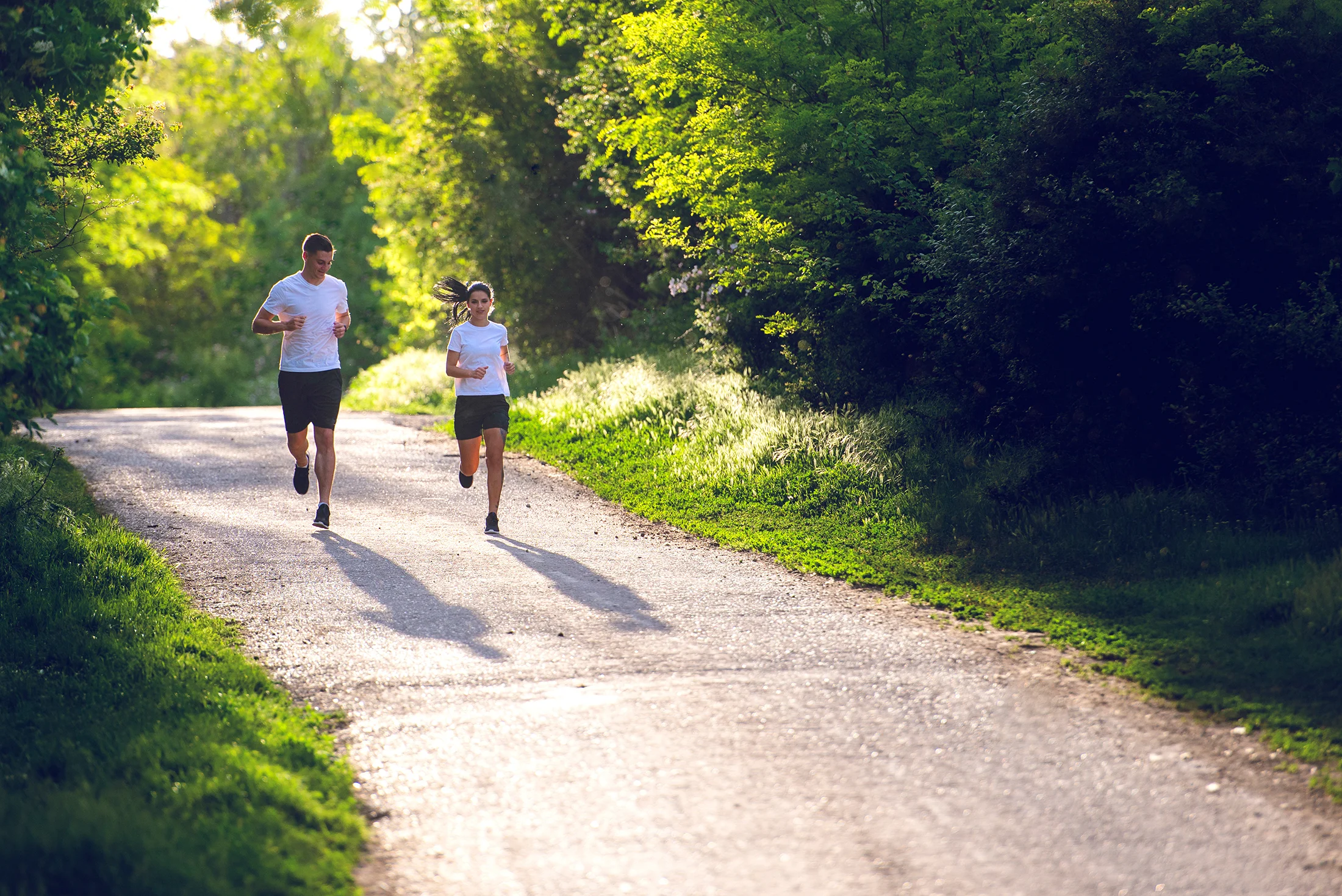 Two people jogging on a sunlit path surrounded by greenery, both wearing white shirts and shorts.