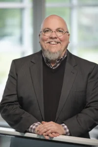 Chad O'Kane, a man with glasses and a grey goatee, wearing a dark suit jacket and sweater vest, stands indoors, smiling at the camera.