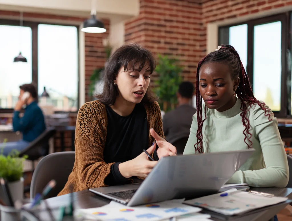Two women sit at a desk in an open office environment, looking at a laptop screen and discussing work.