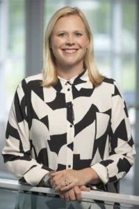 Hayden Shurgar, a woman with blonde hair wearing a black and white patterned blouse stands indoors, smiling with her hands resting on a glass railing.