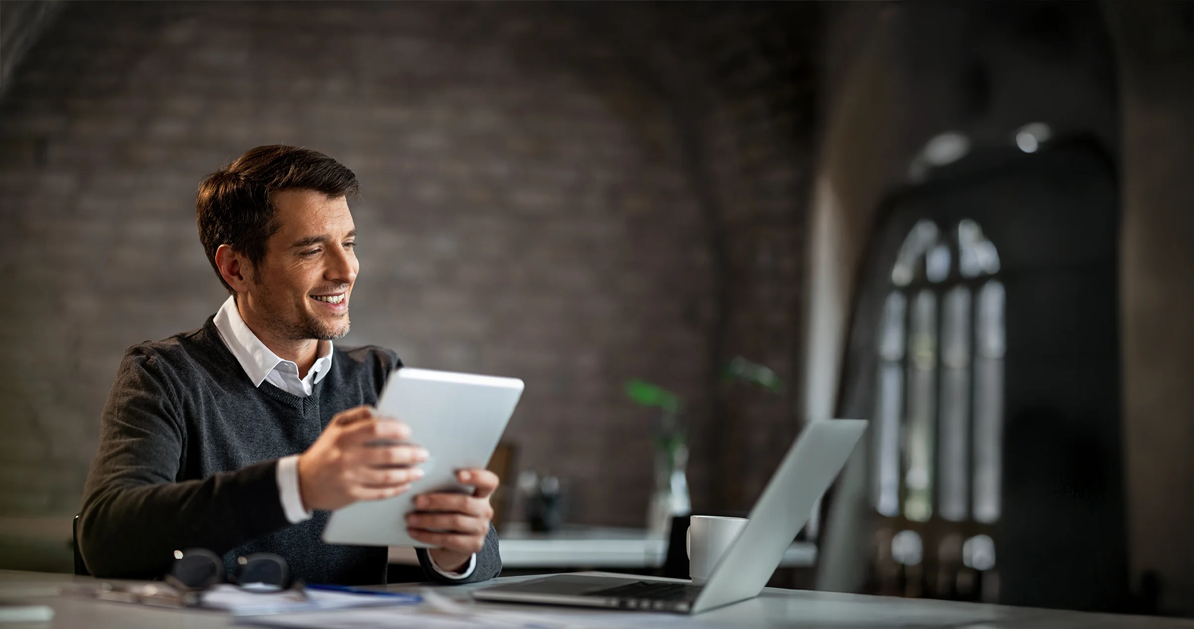 A man sits at a desk in an office, smiling while holding a tablet, with an open laptop, notebook, and coffee cup in front of him—ready to explore new healthcare jobs and EngageMED careers.