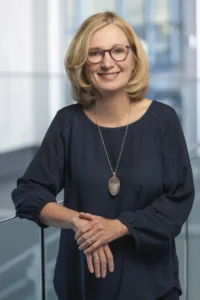 Mary Andrews, a woman with blonde hair and glasses, wearing a navy blouse and pendant necklace, stands indoors, leaning on a railing and smiling at the camera.