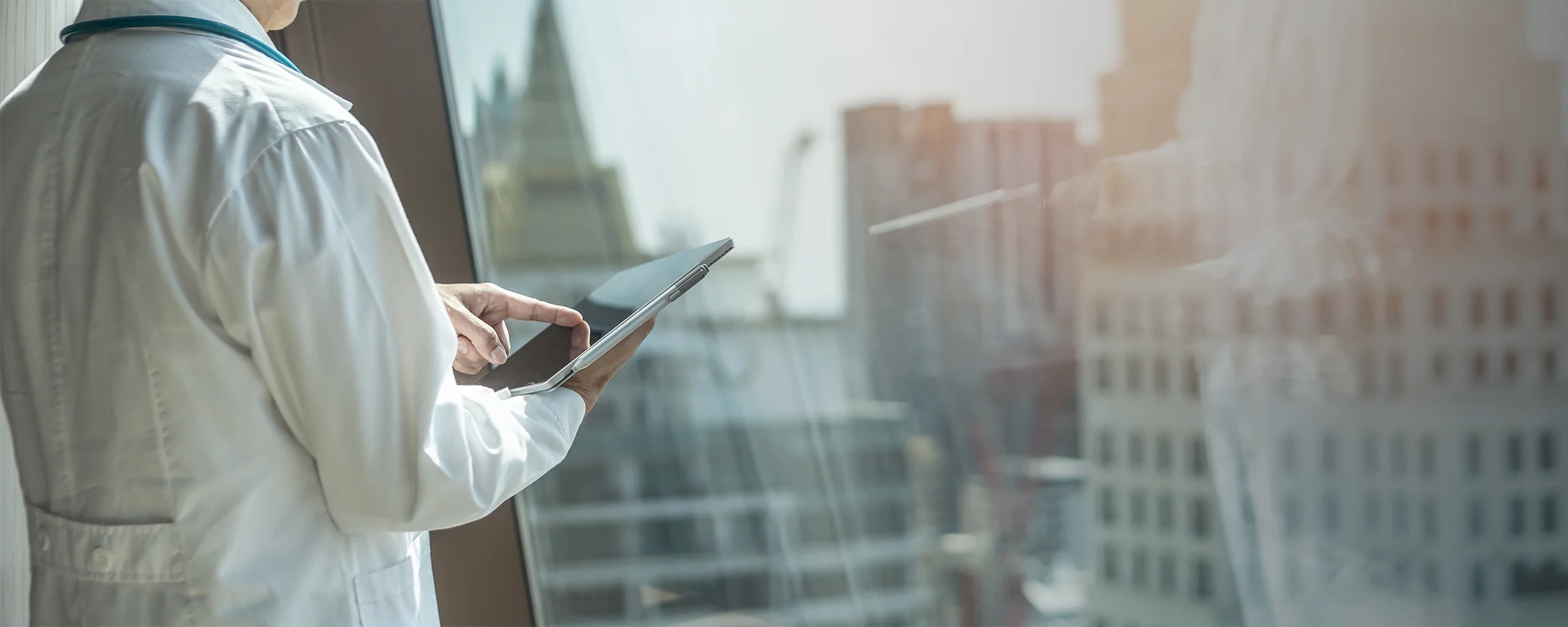 A person in a white lab coat uses a tablet while standing by a large window overlooking a cityscape with tall buildings.