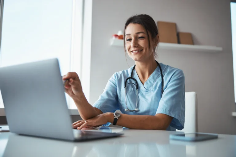 A healthcare professional in scrubs with a stethoscope uses a laptop at a desk in a bright office.