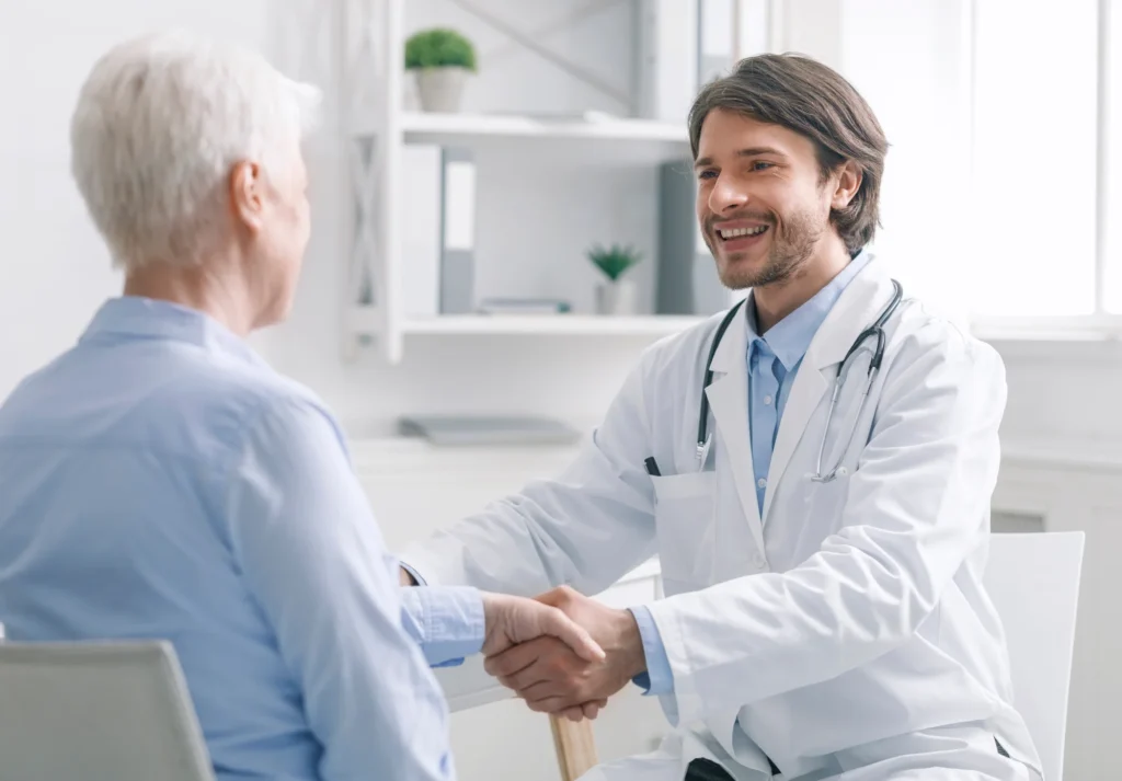A doctor wearing a white coat and stethoscope shakes hands with an older patient in a medical office, highlighting the importance of trust builders in strong patient-provider relationships.