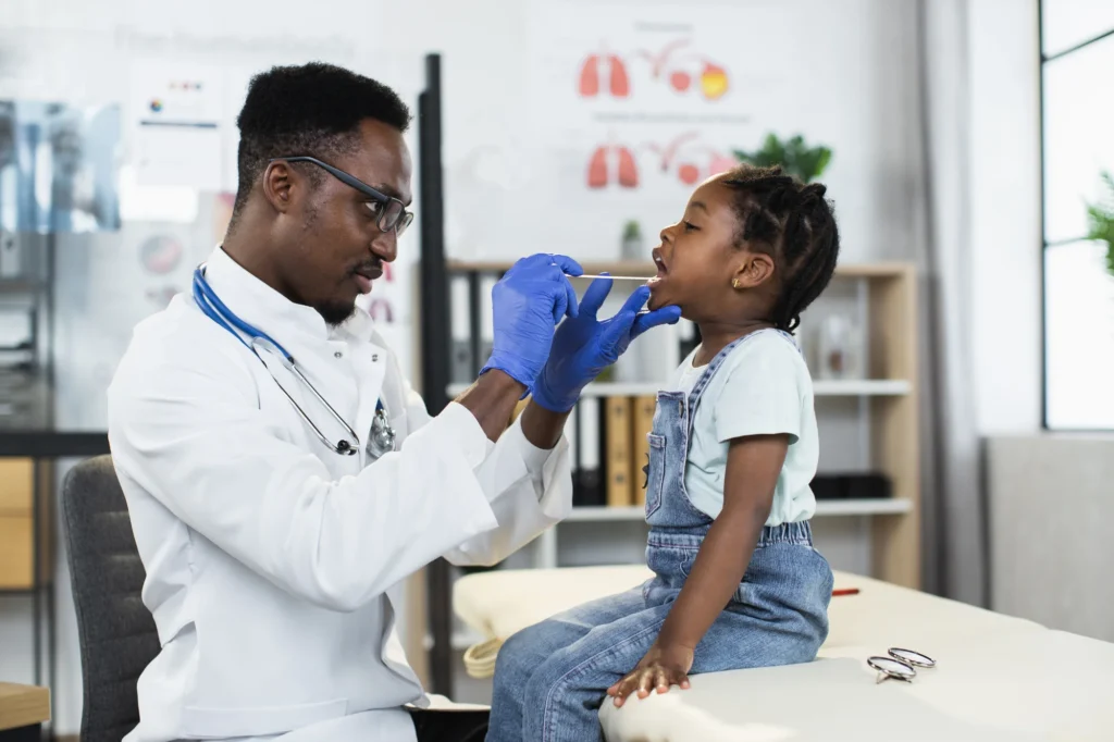 A doctor wearing gloves examines a young child's throat with a tongue depressor in a medical office, showcasing the attentive care provided by Primary Care Providers.