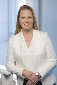 Tiffany Hogan stands indoors near a railing, smiling and wearing a white blazer.