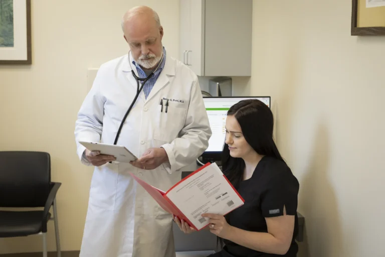 A doctor in a white coat reviews a tablet while a healthcare professional in black scrubs looks at a red folder in a medical office.