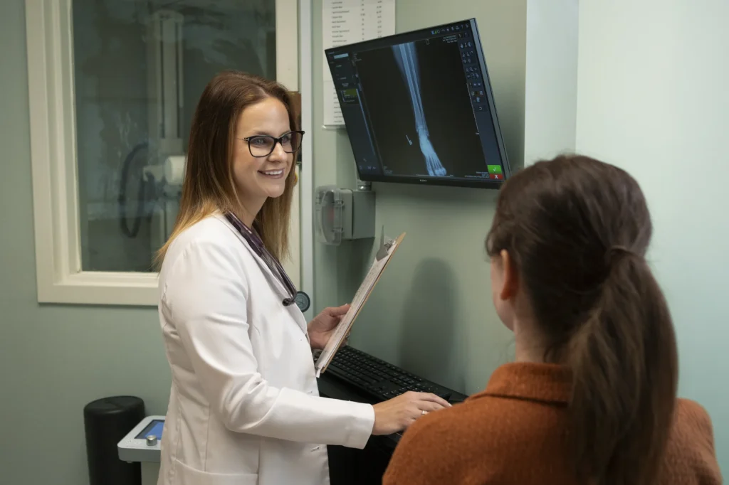 A doctor holding a clipboard discusses an X-ray with a patient in an exam room; an X-ray image is displayed on the monitor.