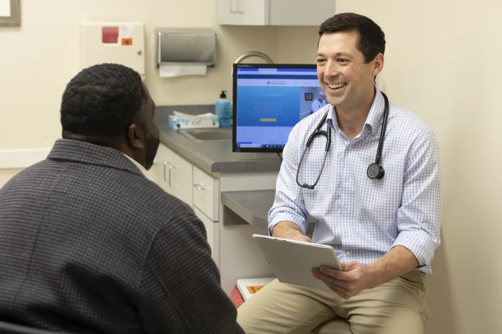 A doctor holding a tablet smiles while talking to a patient in a medical office. A computer screen and medical supplies are visible in the background.