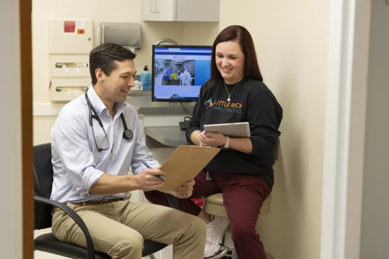 A male doctor and a female healthcare worker review patient information together in a medical office, with a computer monitor in the background.