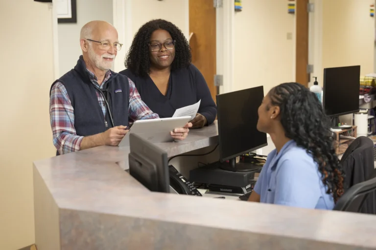 Two people stand at a reception desk speaking with a seated receptionist in a medical office setting.