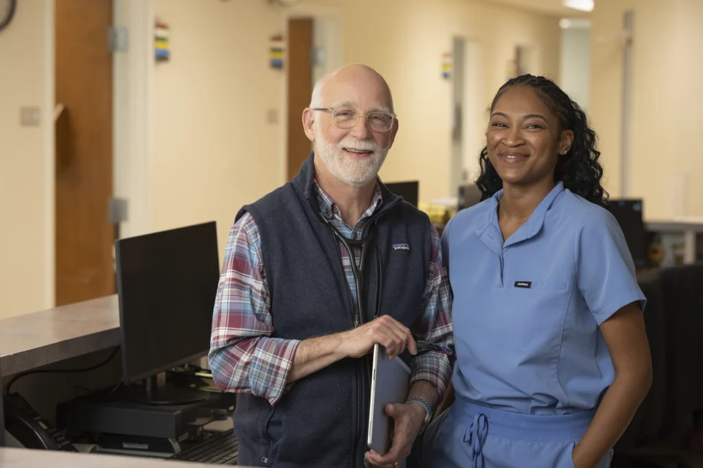 An older man in casual business attire and a woman in medical scrubs stand smiling together in a professional office or clinic setting.