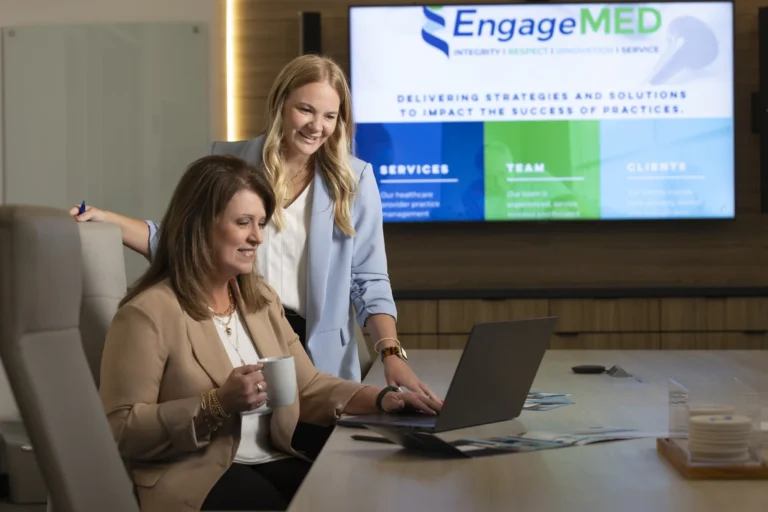 Two women in business attire work together at a laptop in a conference room. A presentation slide for EngageMED is visible on a large screen in the background.