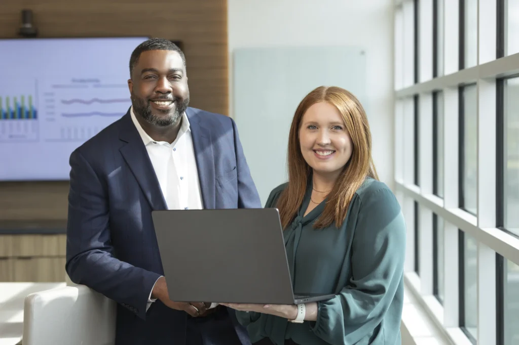 Two professionals standing indoors, one holding an open laptop, both smiling at the camera. A presentation screen with charts is visible in the background.