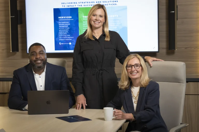 Three business professionals, two women and one man, pose together in a conference room, with a laptop and documents on the table and a presentation screen in the background.