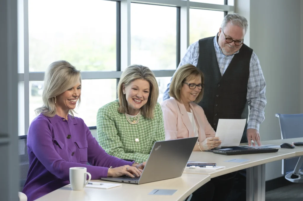 Four adults in business attire sit and stand around a conference table with laptops and papers, smiling and working together in a modern office setting.