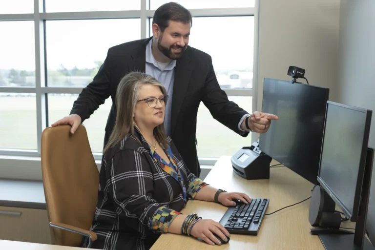 A woman sits at a desk using a computer while a man stands beside her, pointing at the monitor. Both are in an office with large windows in the background.