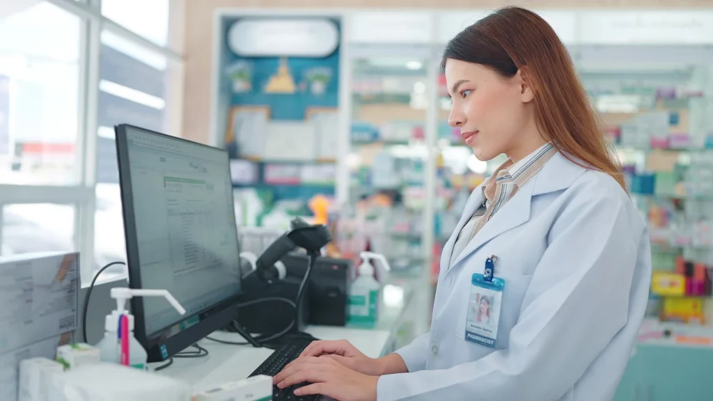 A pharmacist in a white coat works at a computer behind the counter in a pharmacy, ensuring healthcare compliance, with shelves of medication visible in the background.